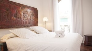Ein ordentlich bezogenes Bett mit weißen Leinentüchern, ein dekoratives Holz-Kopfbrett, eine Lampe und ein Blick ins Freie durch ein Fenster mit Vorhängen.