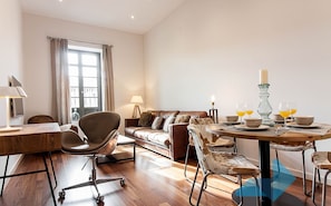 A modern living room with a wooden dining table, leather sofa, and a large window with curtains.