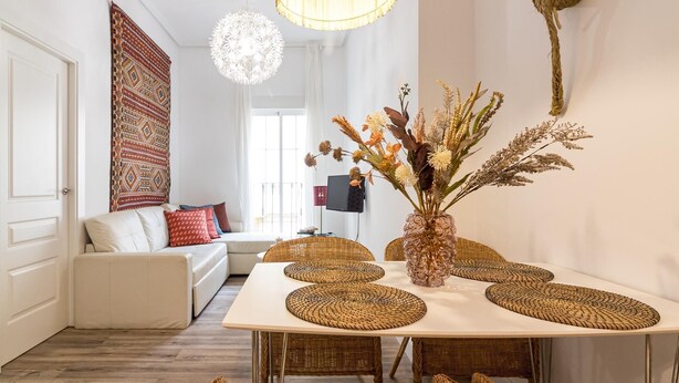 Ein modernes Wohnzimmer mit einem weißen Sofa, einem Esstisch mit gewobenen Untersetzern und einer dekorativen Vase mit getrockneten Blumen.