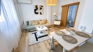 Una sala moderna con sofá, mesa de centro y mesa de comedor. Hay una lámpara colgante, una silla y una puerta que da a otra habitación.