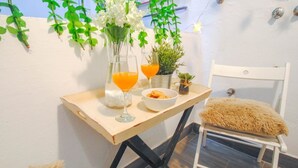 Una mesa pequeña con una silla blanca, un jarrón con flores blancas, un vaso de jugo de naranja, un tazón de bocadillos y una pequeña planta.