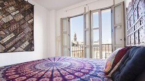 Quarto com cama grande, vista para a cidade através de janelas abertas e tapeçaria na parede.