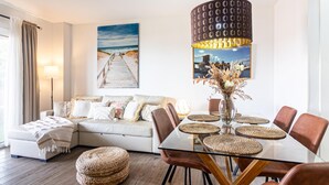 A modern living room with a glass dining table, woven ottoman, and a large painting of a beach. - Malaga Exclusive Beach Apartment with Terrace (Málaga)