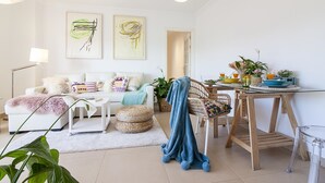 Uma sala de estar moderna com sofá, mesa de centro e área de jantar. Há duas pinturas na parede, um abajur e uma planta.