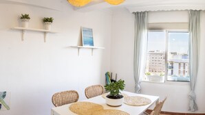 A dining area with a white table, wicker chairs, and woven placemats. There are two potted plants on white shelves, a framed picture on the wall, and a view of buildings through a window with curtains. - Apartamento con Balcon. Free Parking (Málaga)