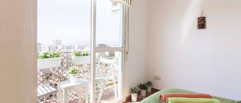 Ein Balkon mit weißen Stühlen und Blumentöpfen, ein Bett mit grüner Bettwäsche und eine Aussicht auf die Stadt durch ein großes Fenster.