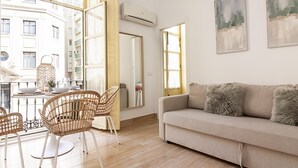 Ein modernes Wohnzimmer mit einem beigefarbenen Sofa, Korbstühlen, einem Esstisch mit Tellern und Gläsern sowie großen Fenstern mit Blick auf ein Gebäude.