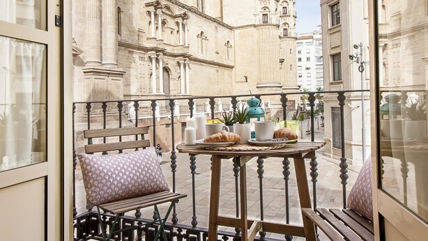 Ein Balkon mit einer hölzernen Bank und einem Tisch, der einen historischen Bau mit aufwendiger Architektur blickt.