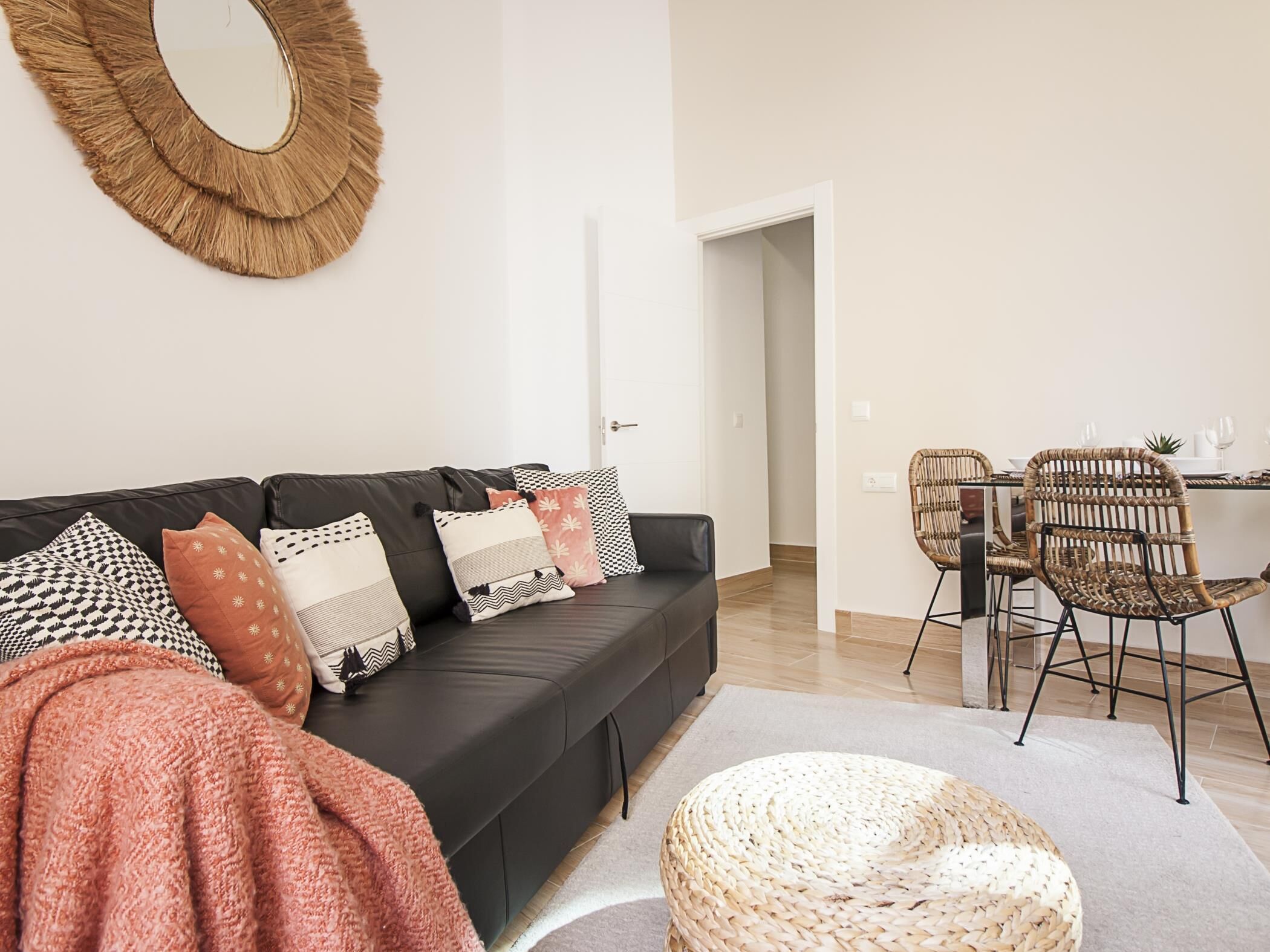 A modern living room with a black sofa, patterned cushions, a woven round mirror, and a dining area with wicker chairs and a table.