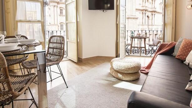 Een kamer met een eettafel gedekt met borden en glazen, rieten stoelen, een bank met kussens, een televisie aan de muur en grote ramen met uitzicht op historische gebouwen.