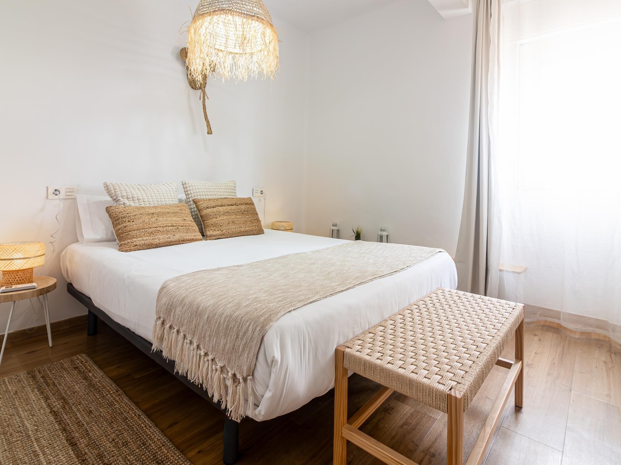 Une chambre à coucher comprenant un lit, des tables de chevet, un banc en osier, un luminaire suspendu et une vue sur l’extérieur par une fenêtre.
