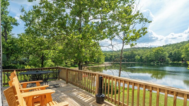 Eine Holzterrasse mit Adirondack-Stühlen und einem Tisch, mit Blick auf einen See, im Hintergrund Bäume und Hügel.