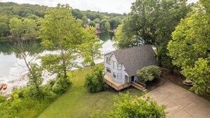 Ein Haus mit einer Terrasse, das auf einen See blickt, umgeben von Bäumen und viel Grün.