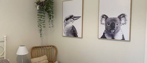 A bedroom with a bed, a wicker chair with cushions, a hanging plant, and two framed animal pictures on the wall.