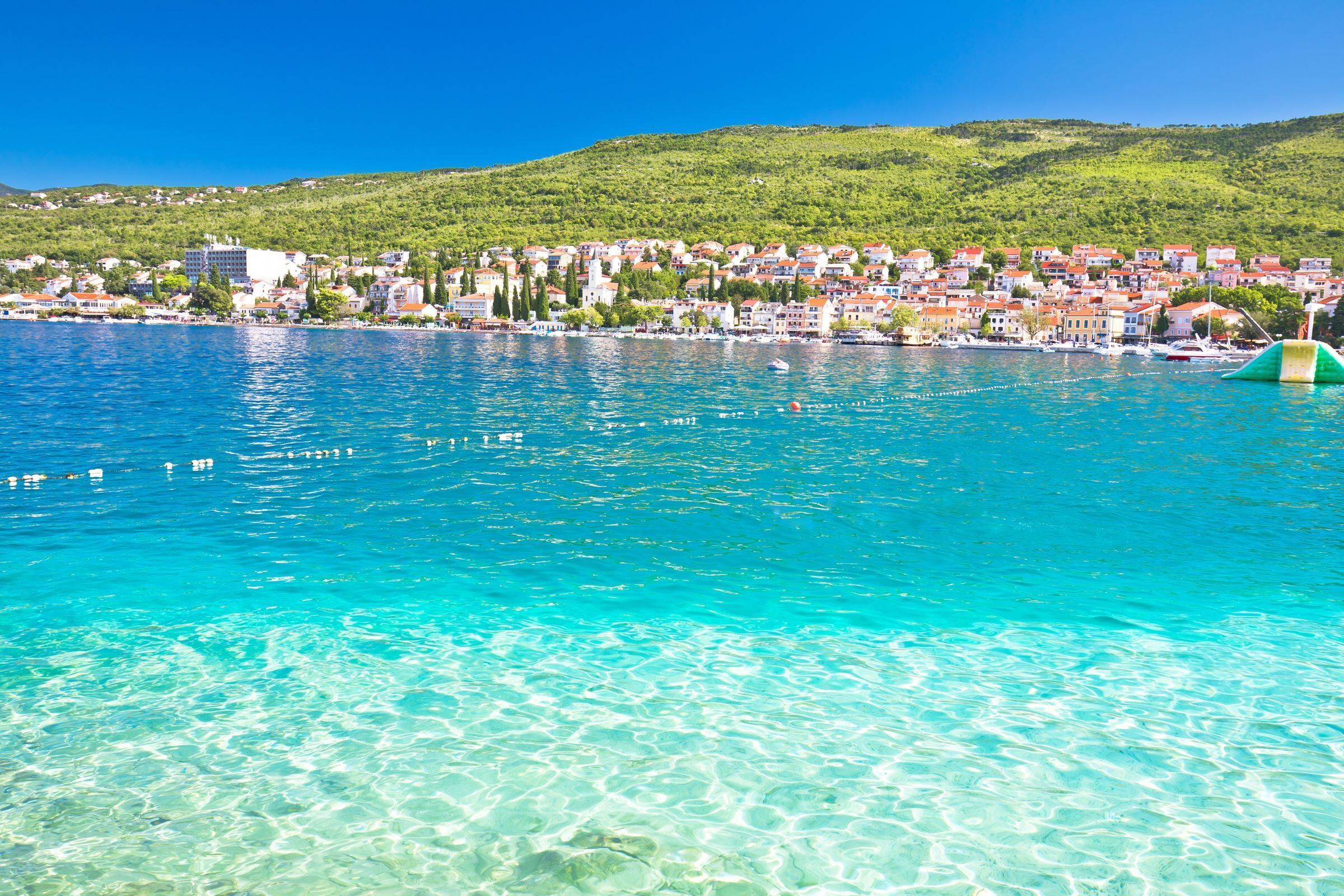 Eine Küstenstadt mit klarem, blauem Wasser, einem grünen Hügel im Hintergrund und einer kleinen, grünen aufblasbaren Struktur im Wasser.