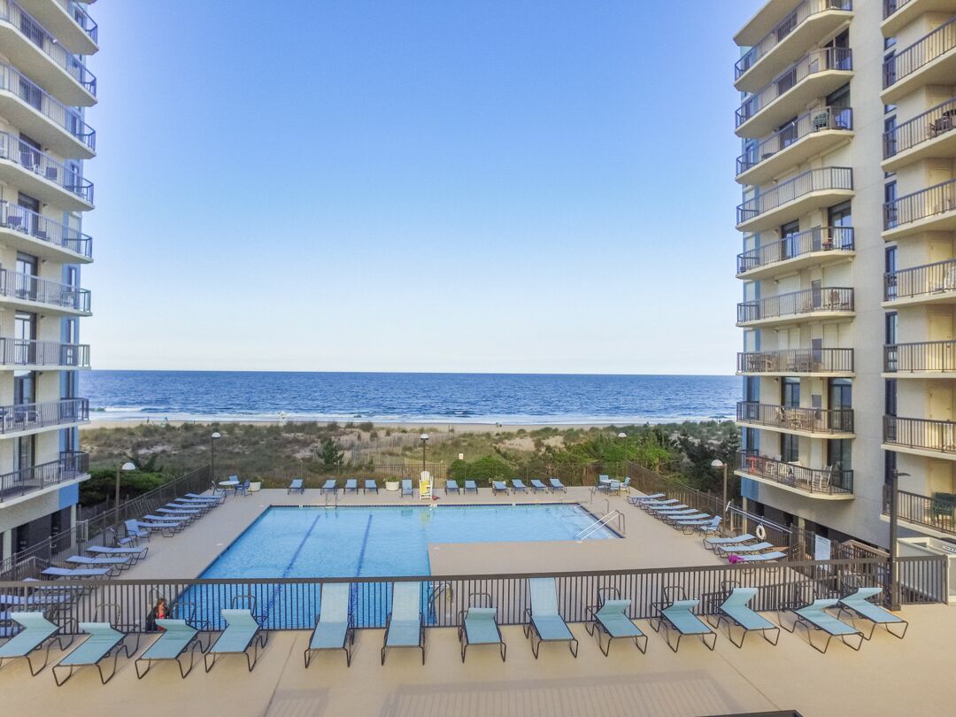 Une piscine sur le toit, dotée de chaises longues, offrant une vue sur la plage et un immeuble de grande hauteur avec des balcons.