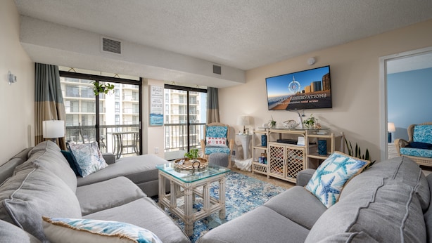 A living room with grey sofas, a glass coffee table, and a beach-themed chair. There is a large window with curtains, a television, and a framed picture on the wall. - 2 Bedroom, 2 Bath Side Unit with fantastic views of the ocean and pool (Ocean City)