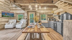 Een rustiek houten interieur met een eettafel, twee geruite fauteuils, een kroonluchter en een keukenhoek.