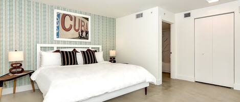 A neatly arranged bedroom with a large bed, a bedside table with a lamp, a wall-mounted artwork, and a wardrobe.