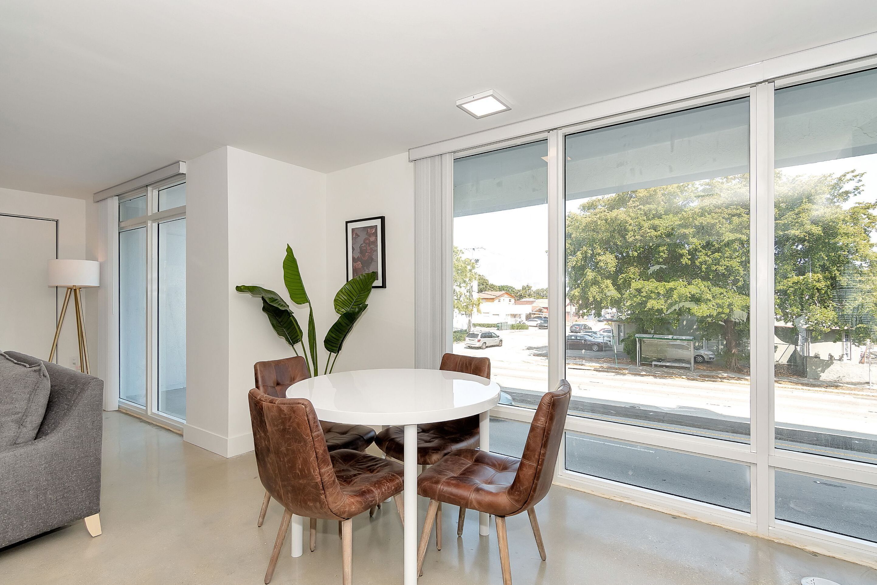 Een moderne woonkamer met een witte ronde tafel, bruine leren stoelen, een grijze bank en grote ramen met uitzicht op de straat en bomen.