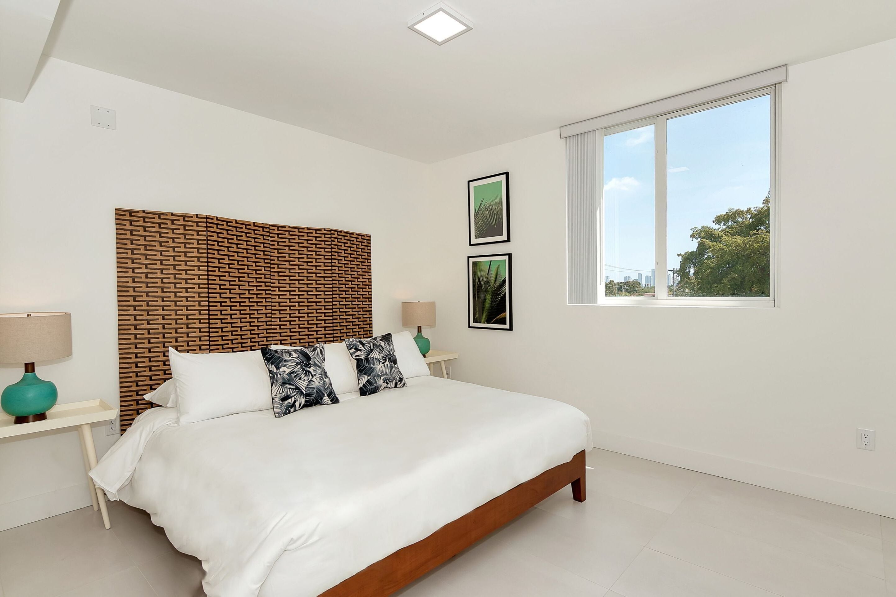 A modern bedroom with a wooden headboard, a large bed with white bedding, two bedside tables with lamps, and a large window offering a view of the outdoors.
