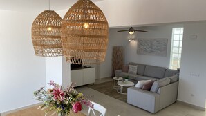 A modern living room with a dining area, featuring a wooden dining table, white chairs, a grey sofa, and a ceiling fan.