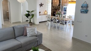 A modern living room with a grey sofa, a dining area with a wooden table and white chairs, a potted plant, and a decorative wall piece.