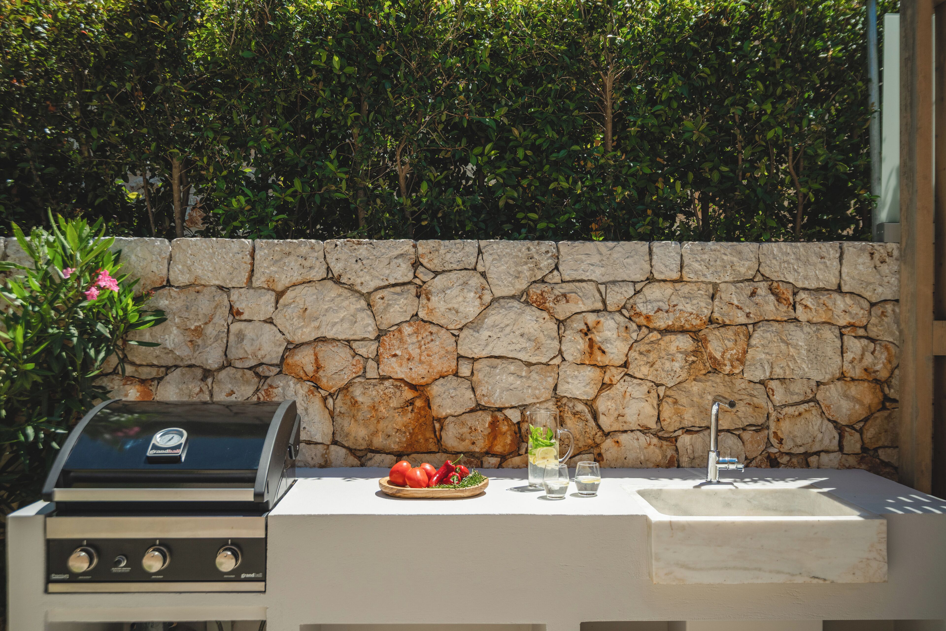 An outdoor cooking area with a stone wall, a grill, and a sink with glasses and a bowl of tomatoes.