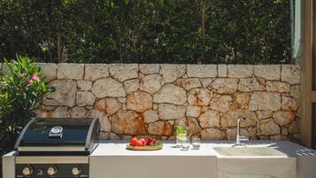 An outdoor cooking area with a stone wall, a grill, and a sink with glasses and a bowl of tomatoes.