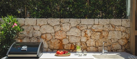 Una zona de cocina al aire libre con pared de piedra, una parrilla y un lavabo con vasos y un bol de tomates.