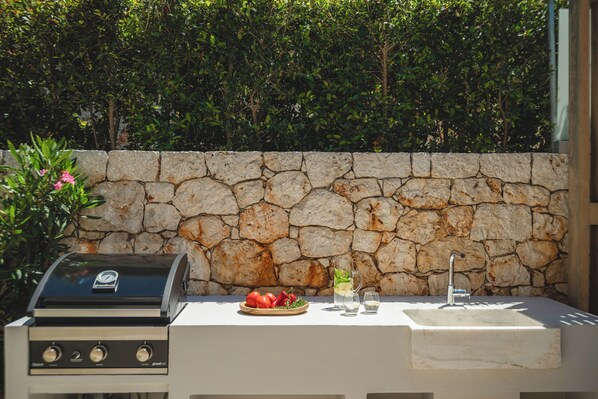 An outdoor cooking area with a stone wall, a grill, and a sink with glasses and a bowl of tomatoes.