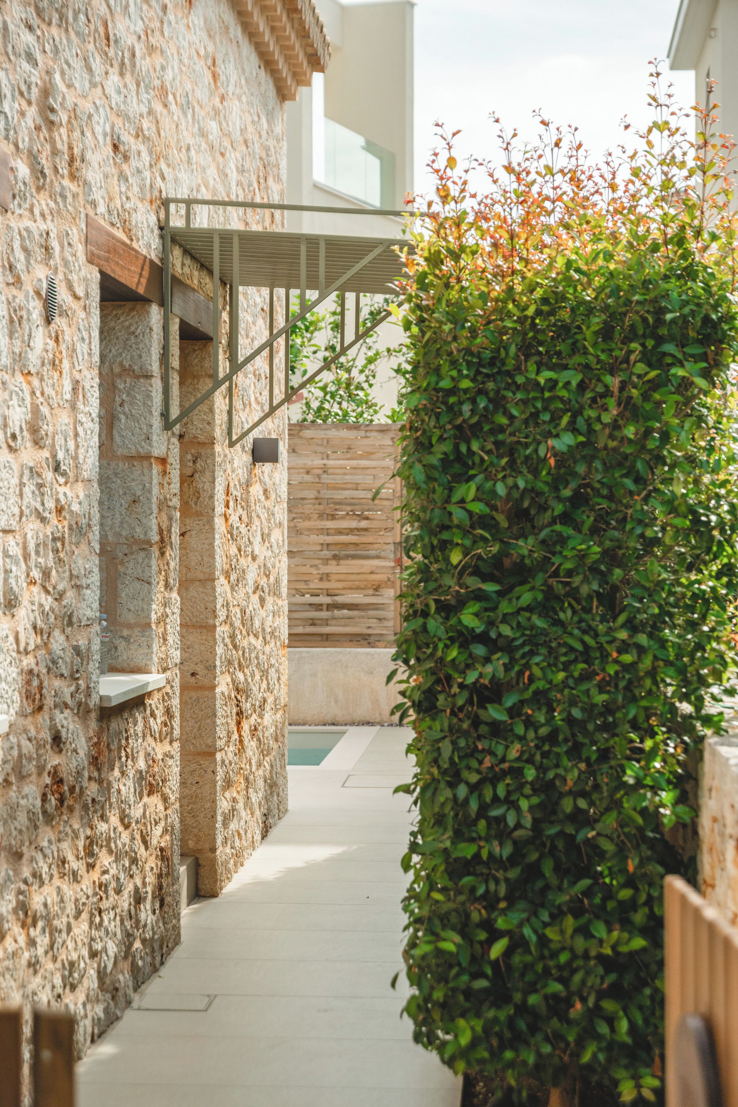 A narrow pathway flanked by stone walls and a wooden fence, leading to a modern building with a metal staircase.