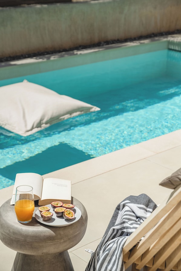 A poolside setting with a lounge chair, a glass of orange juice, a plate of passion fruit, and a book.