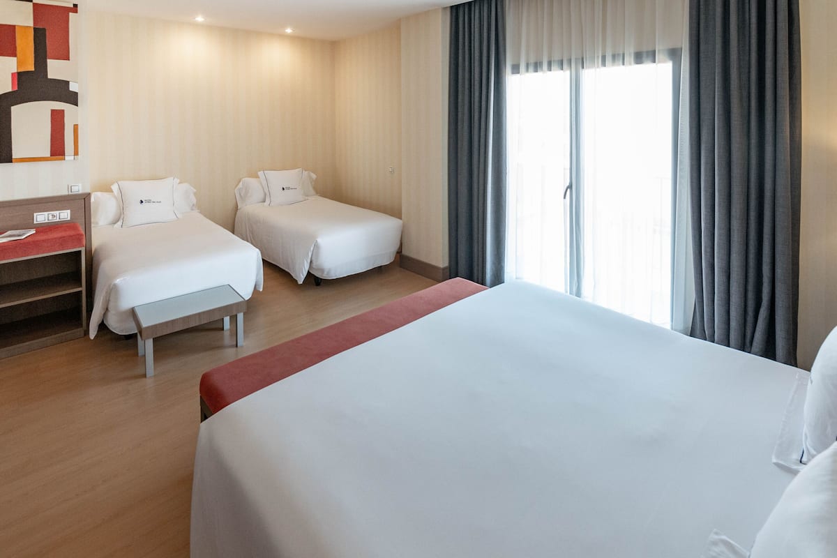 A hotel room with two single beds, a larger bed, a desk with a chair, and a window with curtains.