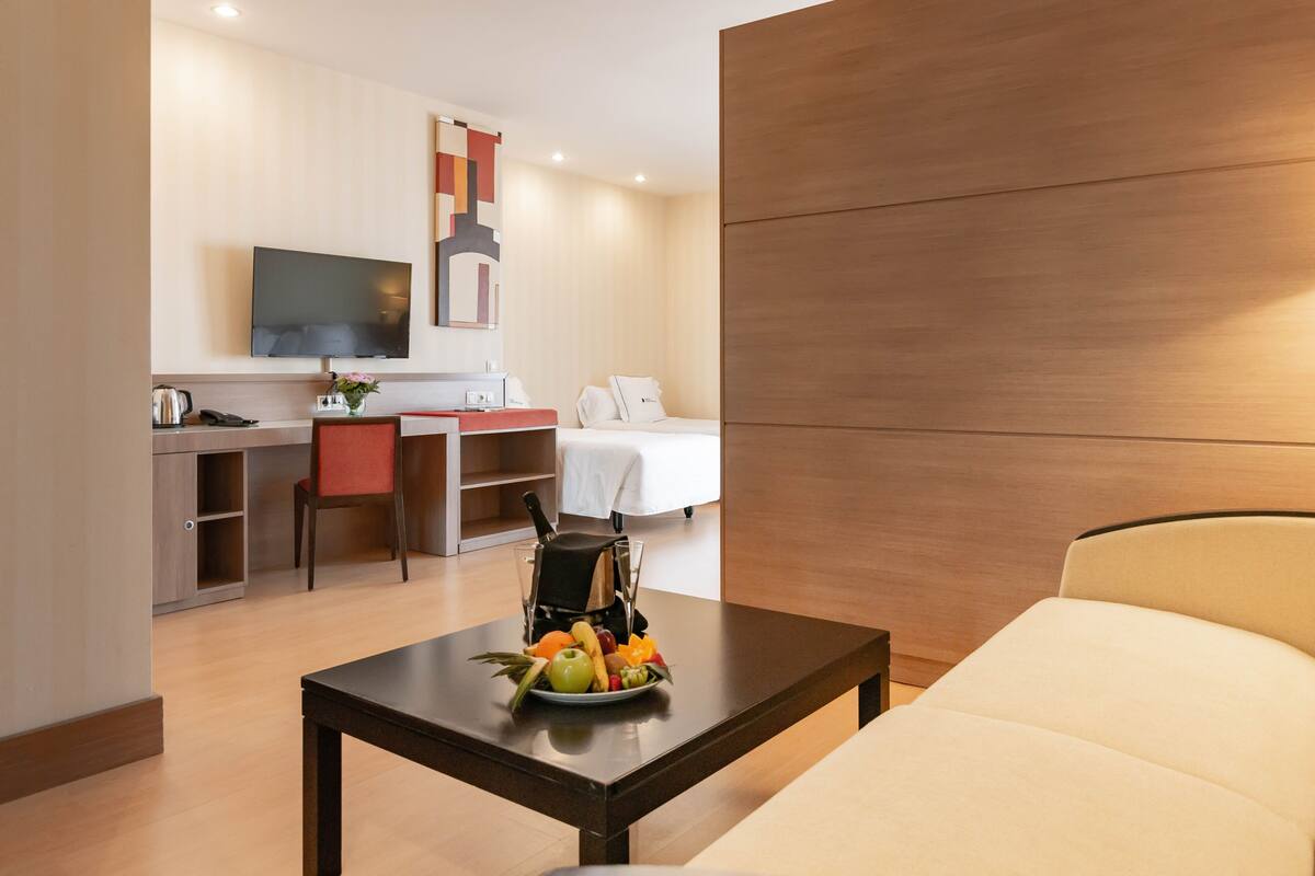 A hotel room with a bed, a desk with a chair, a television, and a small table with fruit.