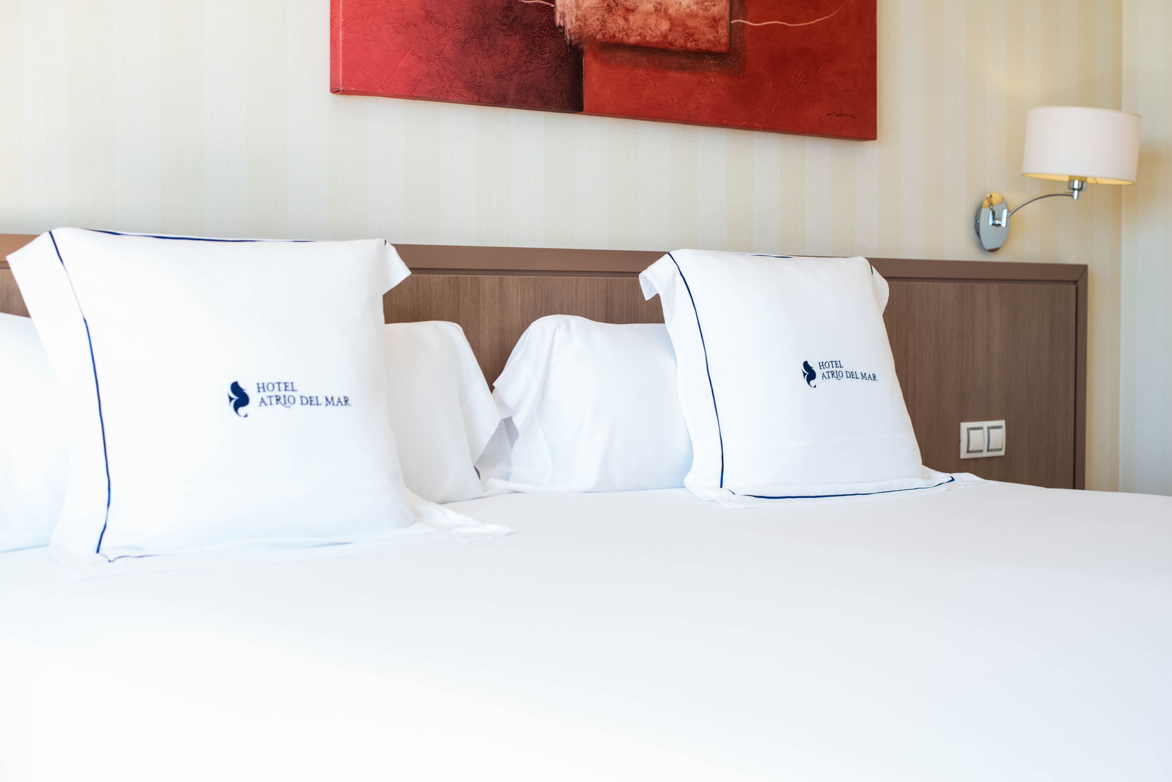 A hotel room with a neatly made bed, pillows with the hotel logo, a wall-mounted lamp, and a red abstract painting.
