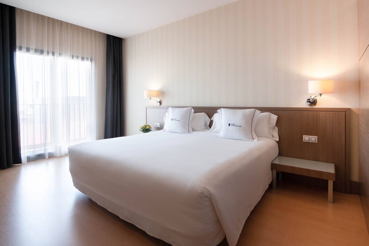 A large bed with white linens, two bedside lamps, a wooden bench, and sheer curtains on a window.