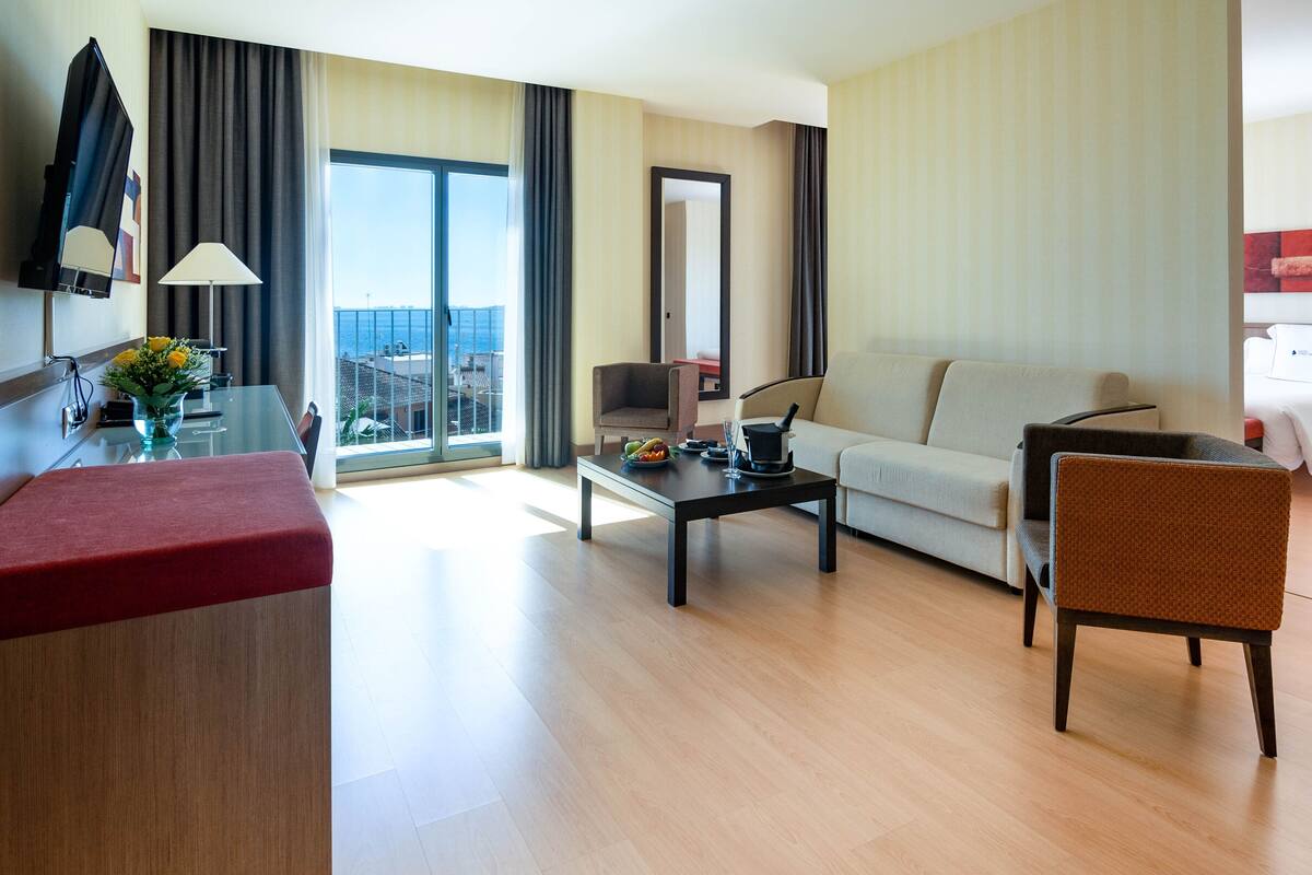A hotel room with a sofa, armchair, dining table, and a large window offering a city view.