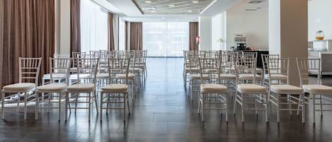 A spacious room with rows of white chairs arranged for seating, large windows with curtains, and a modern ceiling design.