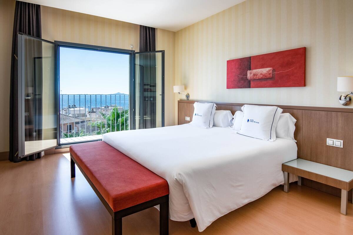 A hotel room with a large bed, a bench, a glass door leading to a balcony, and a red abstract painting on the wall.
