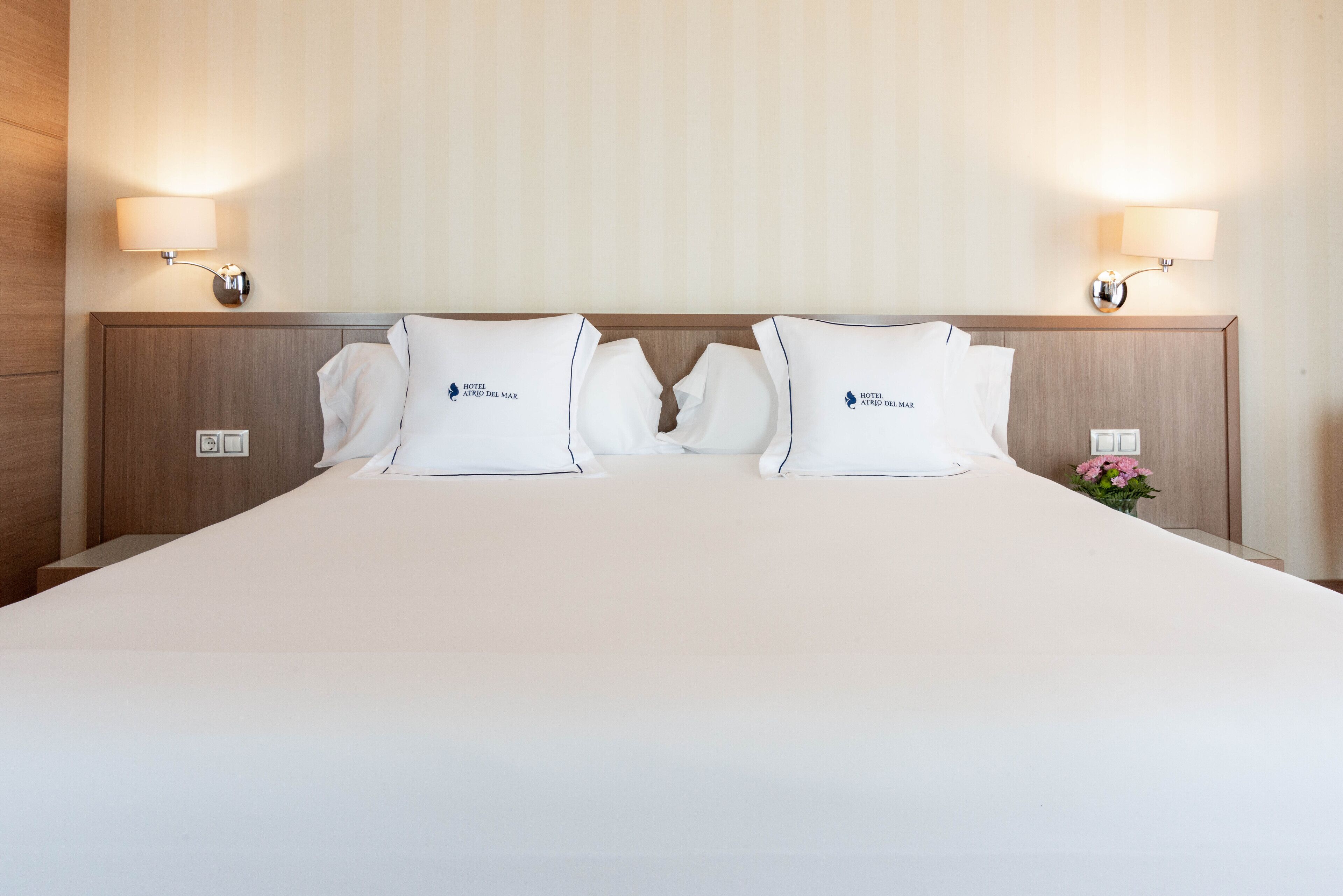 Une chambre d’hôtel avec un grand lit, deux oreillers ornés du logo de l’hôtel, des tables de chevet avec des lampes et un petit vase avec des fleurs.