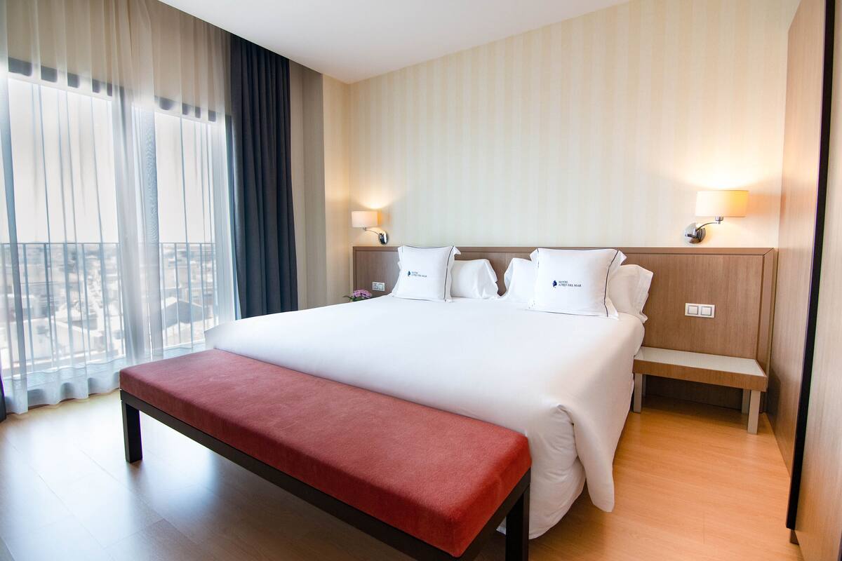 A hotel room with a large bed, a bench, two bedside tables, and a view through sheer curtains.