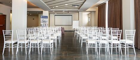 A conference room with rows of white chairs, a projector screen, and a large poster on the wall.