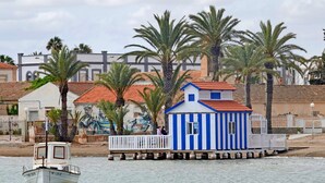 Een klein, blauw-wit gestreept gebouw met een rood dak, op palen boven het water geplaatst, met palmbomen en gebouwen op de achtergrond.