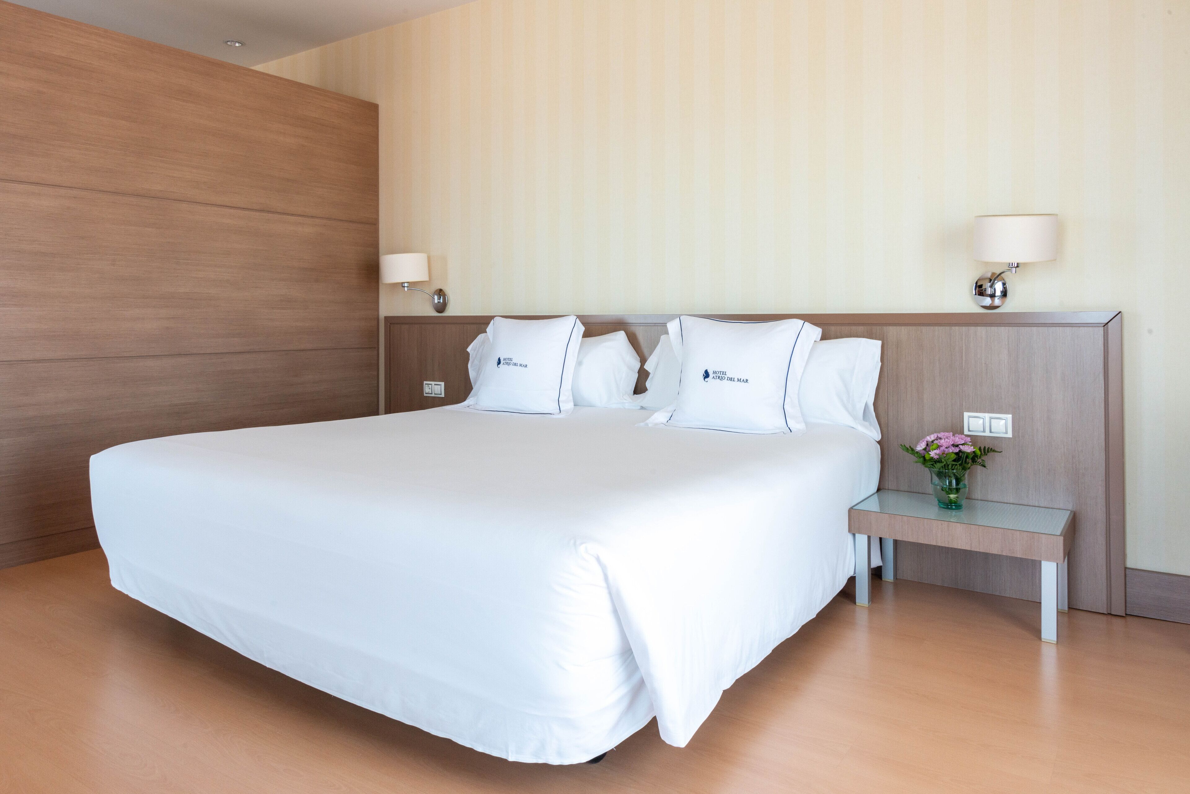 Una cama grande con sábanas blancas, cabecera de madera, dos mesitas de noche con lámparas y una pequeña mesa con un florero.