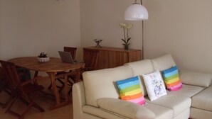 Sala de estar com sofá bege, mesa de jantar de madeira e cadeiras. Há um laptop sobre a mesa, um vaso com flores e uma luminária.