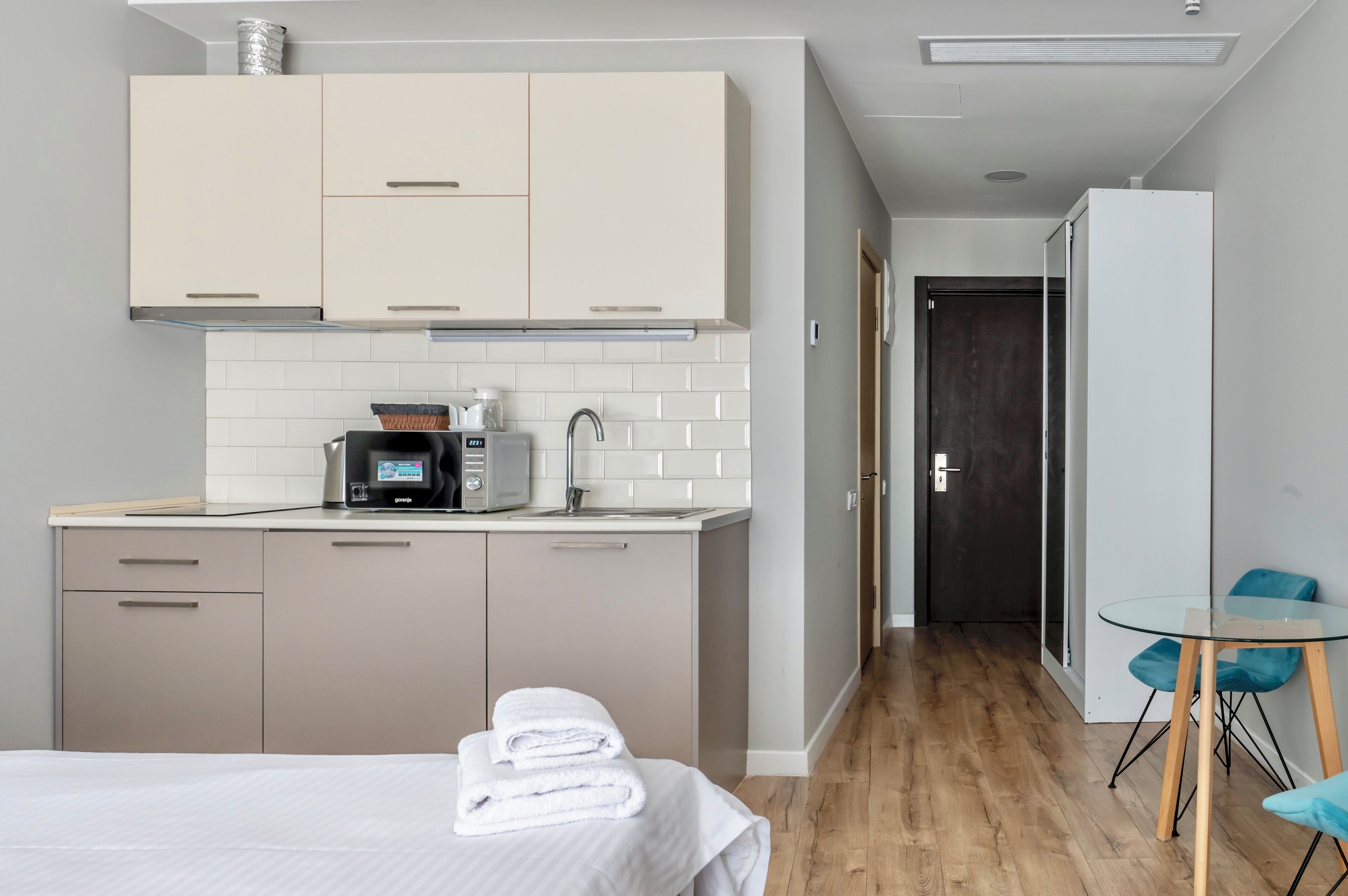 Una habitación de hotel moderna con kitchenette, una cama con toallas ordenadas, una mesa de vidrio con sillas y una puerta oscura al fondo.