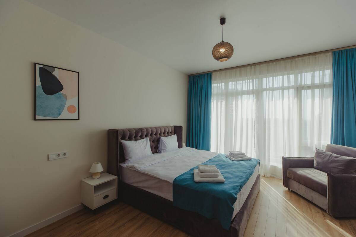 Un dormitorio moderno con cama, mesita de noche, lámpara y vistas al exterior a través de amplias ventanas.