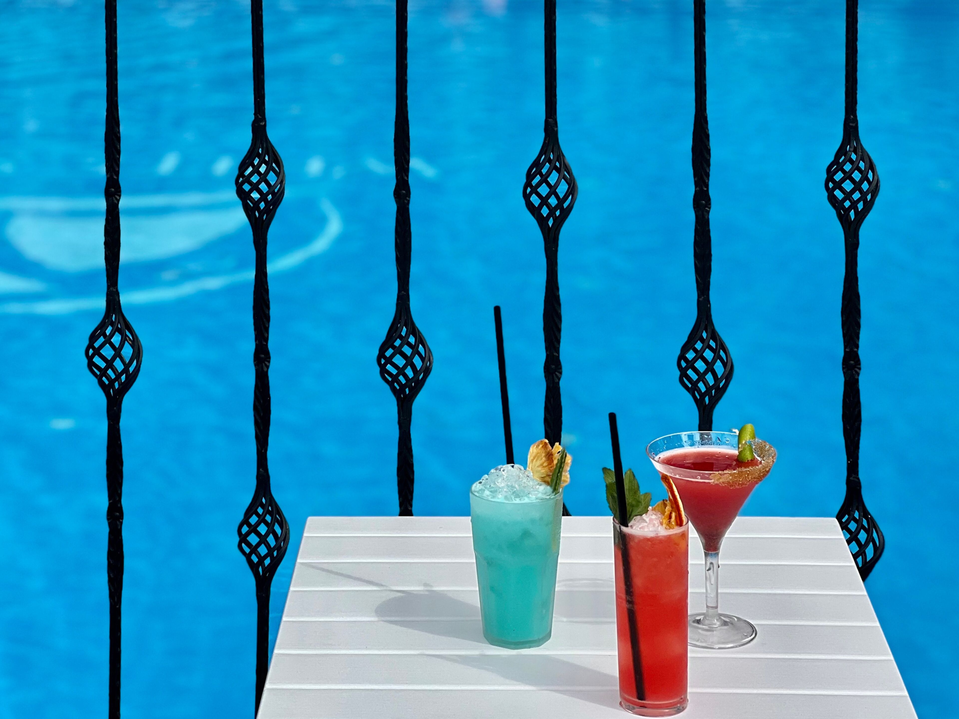 A poolside table with two drinks and a martini glass, set against a blue pool with decorative metal railings.