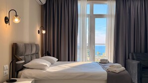 Habitación de hotel con cama, lámparas de noche, mesita de noche, una silla y vistas al mar a través de amplios ventanales.